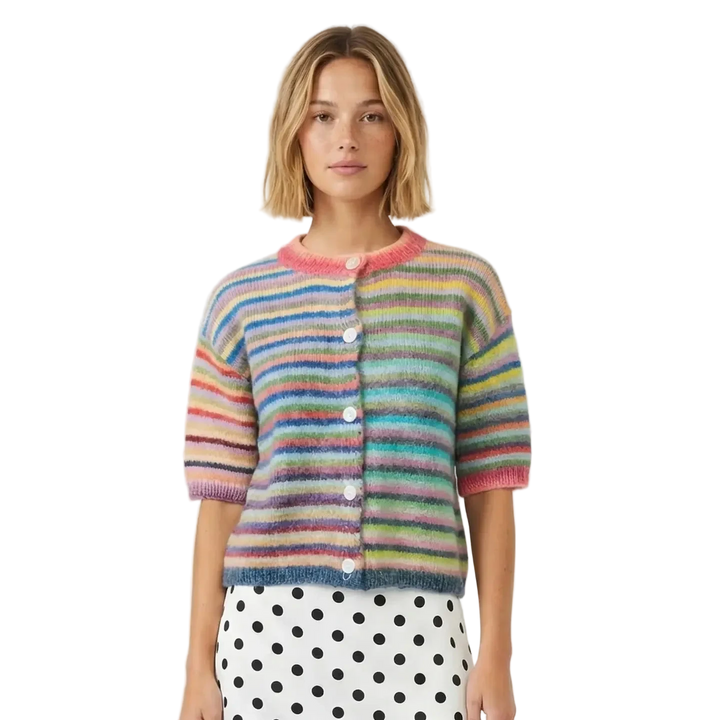 A woman wearing a short-sleeve knit cardigan with horizontal rainbow stripes in pink, blue, yellow, green, and orange, paired with a white polka-dot skirt, against a light gray studio background.