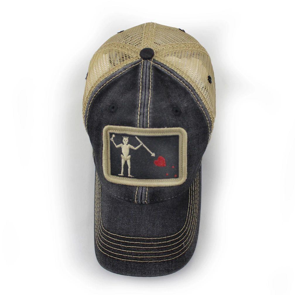 Black trucker hat with embroidered Blackbeard pirate flag patch on the center and khaki mesh back panels. Patch depicts a natural colored skeleton with horns piercing a red bleeding heart with a spear.