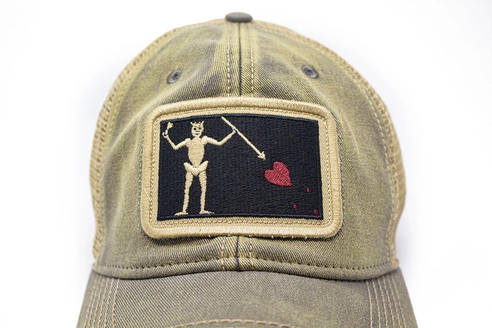 Close up of neutral grey trucker hat with embroidered Blackbeard pirate flag patch on the center and khaki mesh back panels. Patch depicts a natural colored skeleton with horns piercing a red bleeding heart with a spear. 