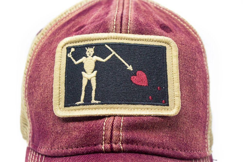 Close-up of brick red trucker hat with embroidered Blackbeard pirate flag patch on the center and khaki mesh back panels. Patch depicts a natural colored skeleton with horns piercing a red bleeding heart with a spear. 