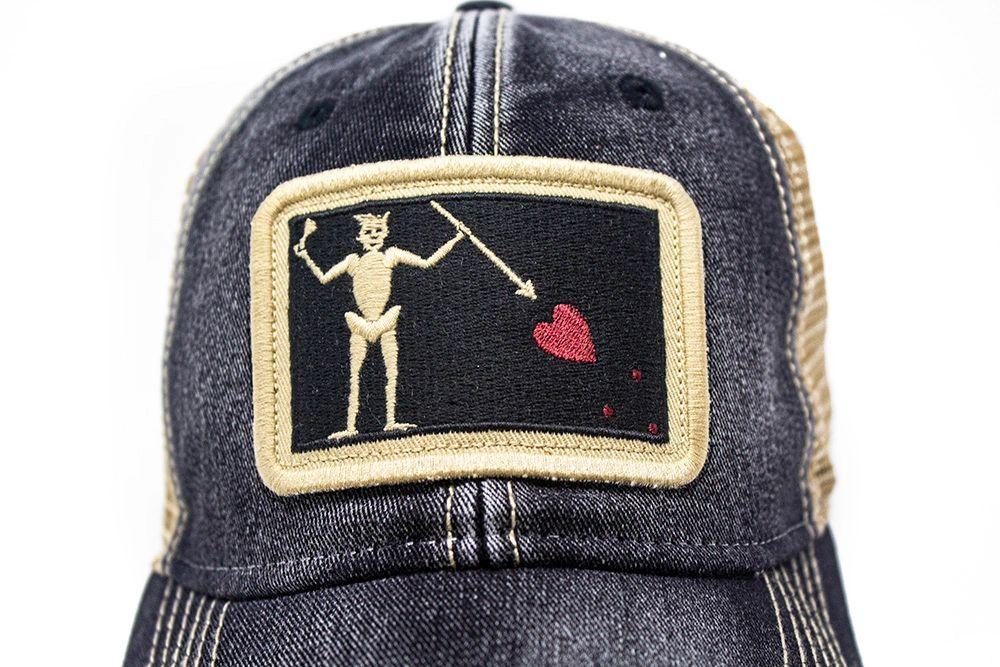 Black trucker hat with embroidered Blackbeard pirate flag patch on the center and khaki mesh back panels. Patch depicts a natural colored skeleton with horns piercing a red bleeding heart with a spear.
